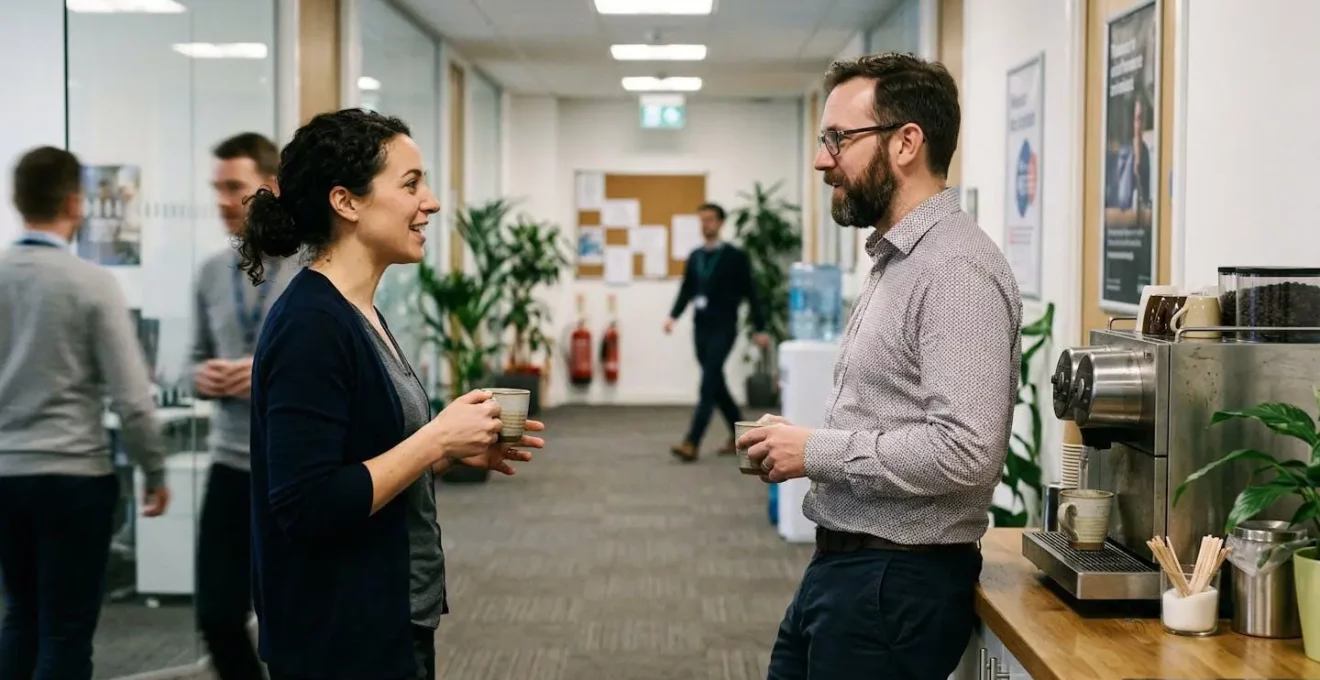 Deux personnes discutant debout près d'une machine à café dans un couloir d'entreprise, ambiance détendue